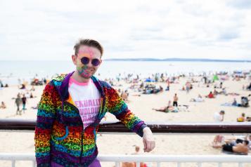 Man stood in front of the beach in a multicoloured top with multicoloured lines painted on his face